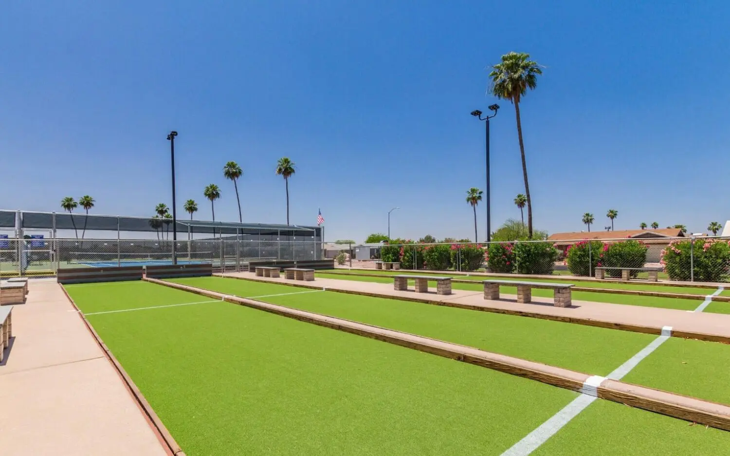 A sunny outdoor sports facility in San Tan Valley features several bocce ball courts with lush green surfaces. Benches line the courts, and palm trees sway gently against a clear blue sky, showcasing the area's new artificial turf installation.