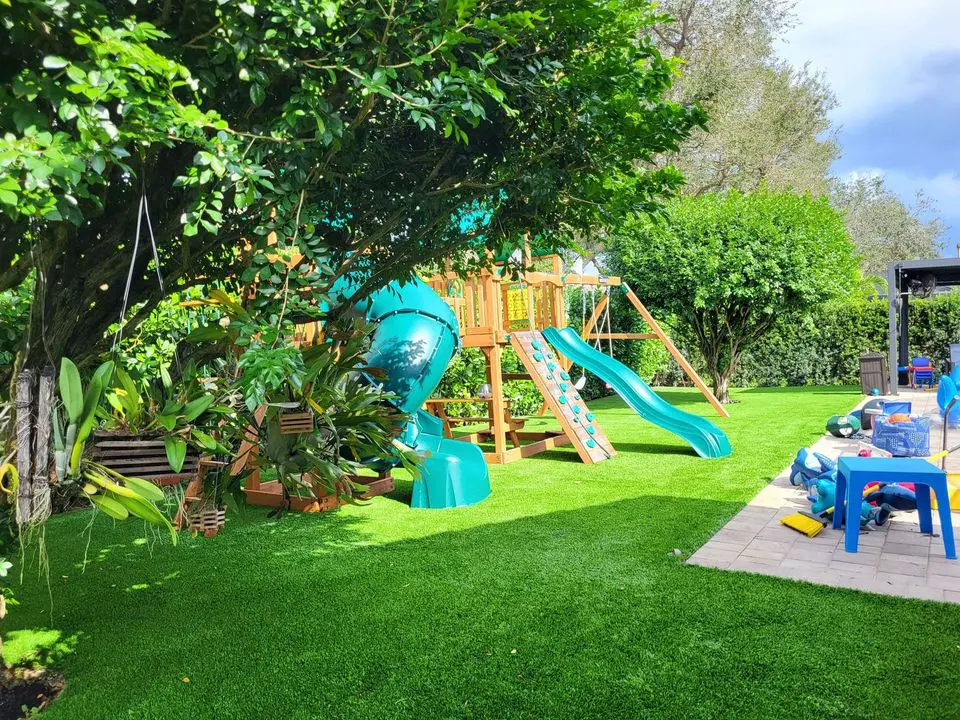 A vibrant backyard in San Tan Valley, with a wooden playset featuring a green slide and climbing wall, is surrounded by lush greenery. A blue table and chairs are on a stone patio to the right, and various toys are scattered on the grass—consider an artificial grass installation for easy maintenance.