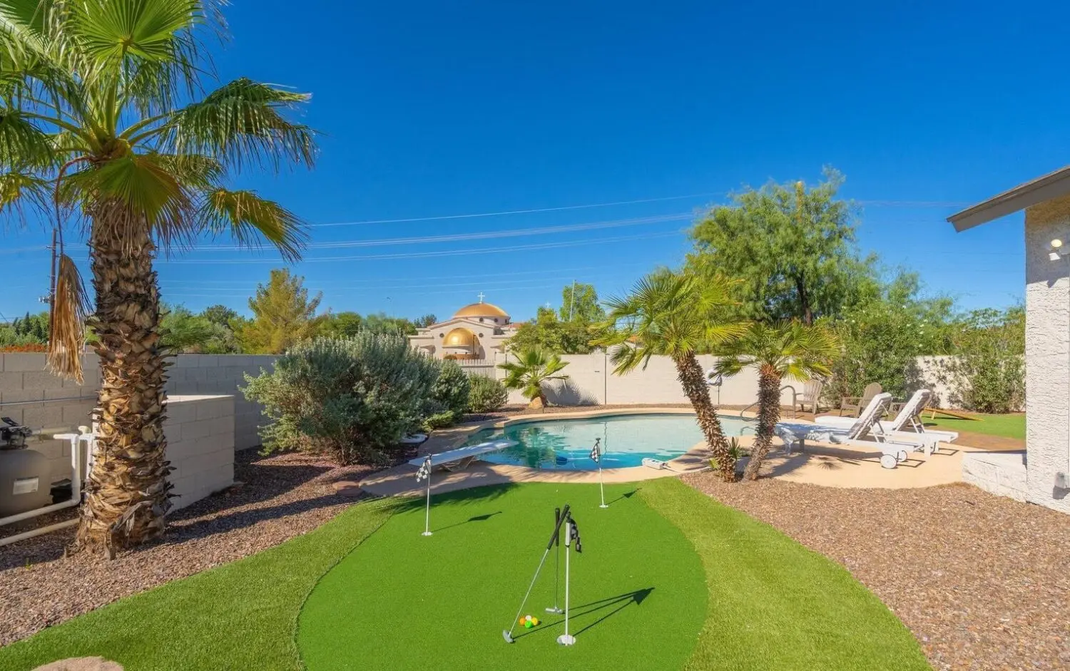 A backyard oasis in San Tan Valley boasts a turquoise swimming pool, surrounded by palm trees and desert landscaping. An inviting putting green with artificial grass graces the foreground, while lounge chairs sit poised on the poolside patio under a clear blue sky.