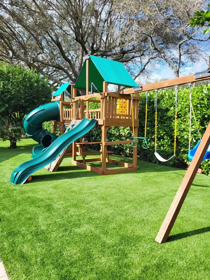 A wooden playground set with a green slide, tube slide, swings, and a canopy roof stands on vibrant artificial grass. Tall trees and bushes surround the play area under a clear blue sky.