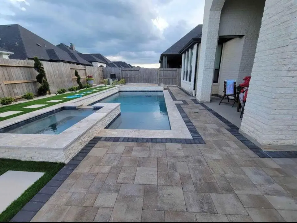 A modern backyard in San Tan Valley, AZ, showcases a rectangular swimming pool with a connected hot tub. The outdoor space is beautifully enhanced with stone tiles and artificial grass. A fence, large house, and cloudy sky create a serene backdrop for this stunning area.