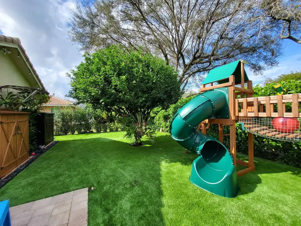 A backyard in San Tan Valley features a lush green lawn with a tree and a wooden play structure boasting a green spiral slide. Encircled by a wooden fence and dense foliage, it offers a serene outdoor play environment perfect for family fun.