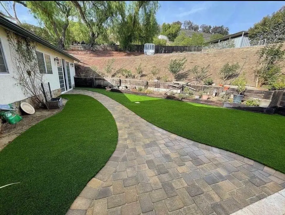 A backyard features a paved stone walkway curving through installations of artificial grass. A garden with plants and a greenhouse is in the background, next to a white house on the left. Sloped land with trees encloses the yard.