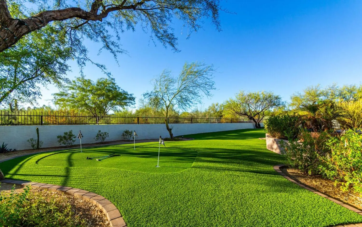 A lush backyard in San Tan Valley features a green artificial turf putting green with several small flags. It's surrounded by desert landscaping, including trees and shrubs, all enclosed by a white fence. The sky above is clear and blue, creating a serene oasis.