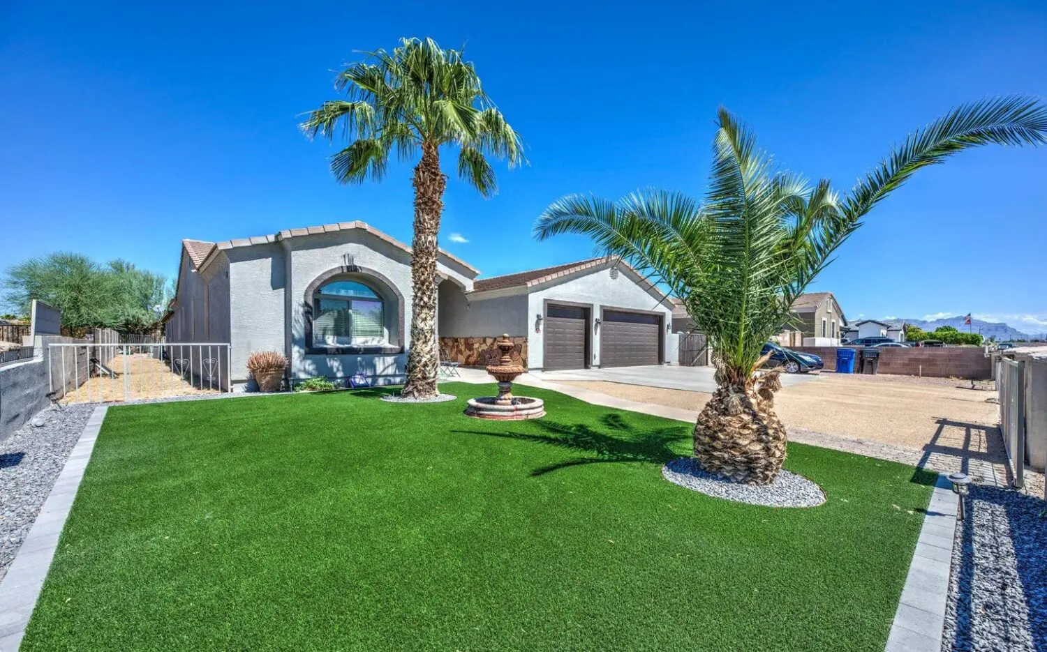 A modern house in San Tan Valley boasts a gray facade and arched entryway, framed by two palm trees and manicured artificial grass from top-tier turf products. The driveway leads to three brown garage doors, all set against a clear blue sky.