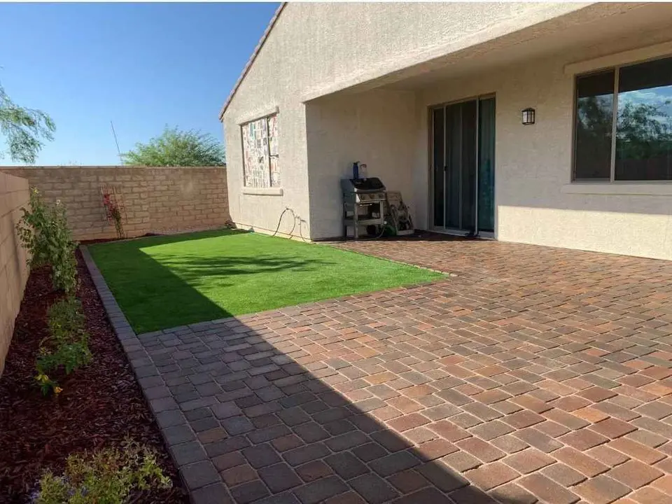 A backyard patio features a concrete brick floor and a rectangular patch of lush artificial grass, complemented by a barbecue grill under a covered area. The yard, adorned with small flowering plants, is bordered by a stone wall, showcasing quality turf products.