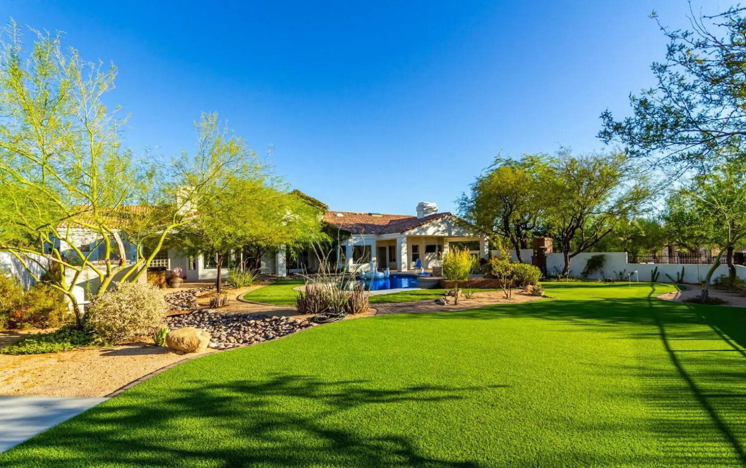 A spacious backyard in San Tan Valley boasts lush green grass, desert landscaping, and a clear blue pool. In the background, a white house with a terracotta roof is framed by various trees and shrubs under the bright blue sky.