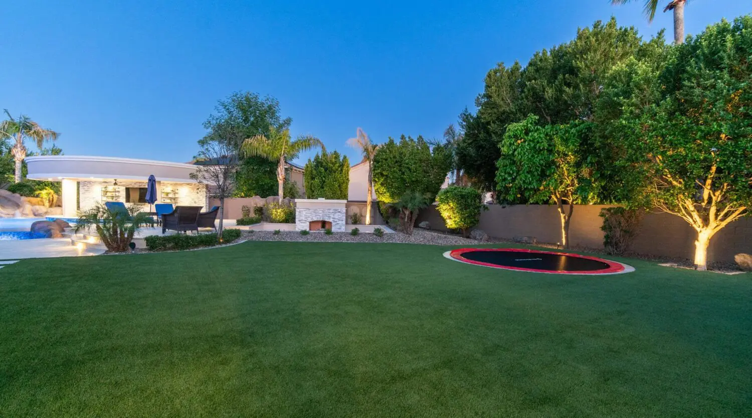 A spacious backyard featuring a well-manicured lawn with a built-in trampoline and premium turf products. The area is surrounded by tall green trees and shrubs. A swimming pool and poolside seating are visible in the background under a clear blue sky.