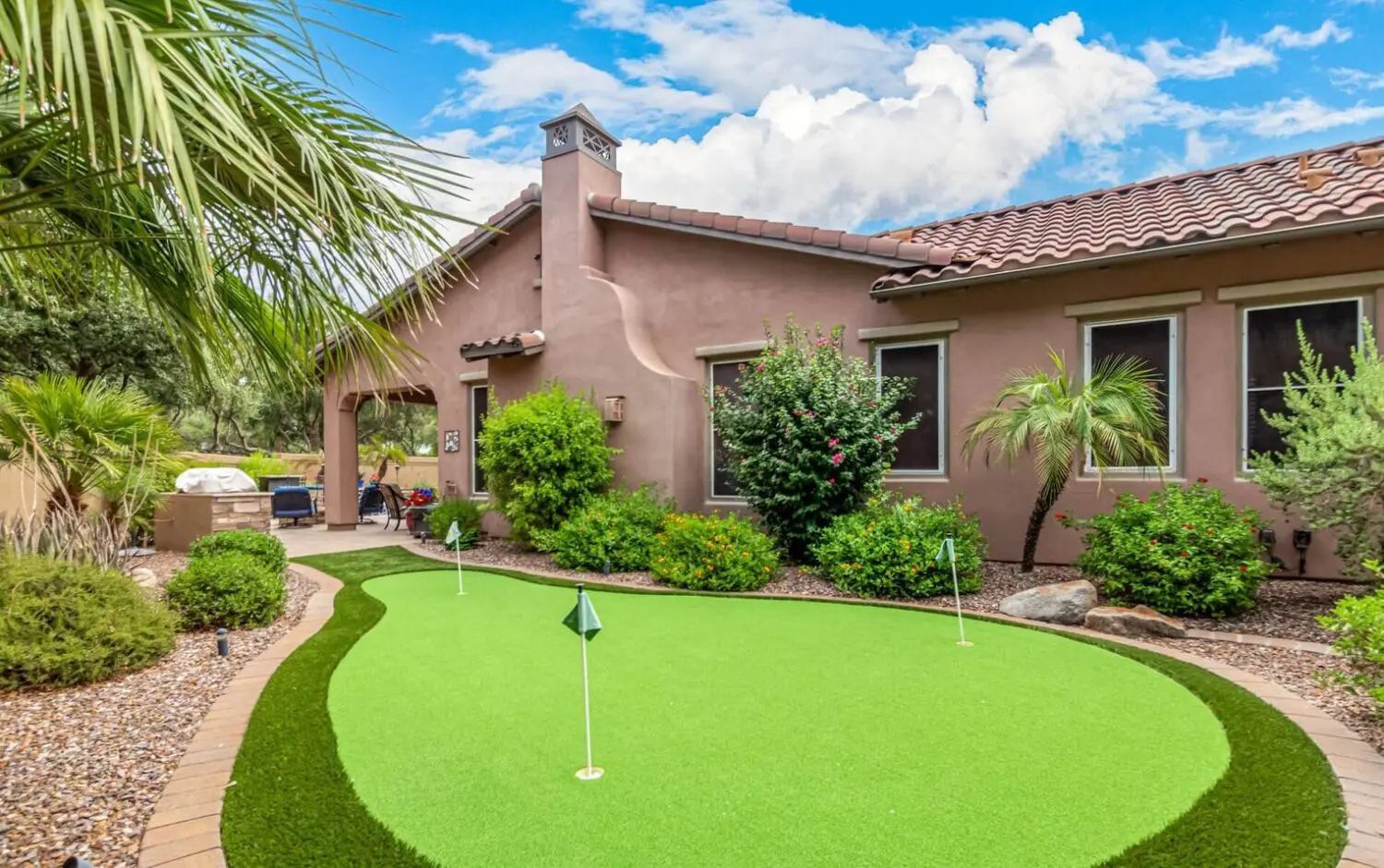 A backyard in San Tan Valley features a small artificial putting green crafted from premium turf products, surrounded by lush landscaping with shrubs, palm trees, and rocks. The house boasts tan stucco walls and a red tile roof, while a barbecue area sits under the cloudy blue sky.