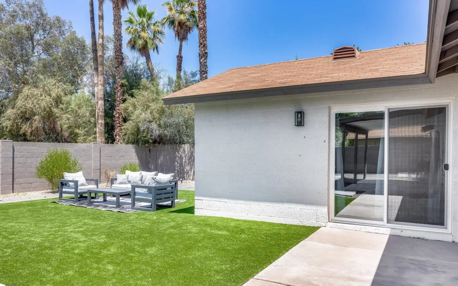 A backyard in San Tan Valley features artificial grass, a white and gray outdoor seating area on a patterned rug, all enclosed by a sleek gray brick wall. Tall palm trees and lush greenery add to the charm, with a house featuring sliding glass doors gracefully positioned to the right.