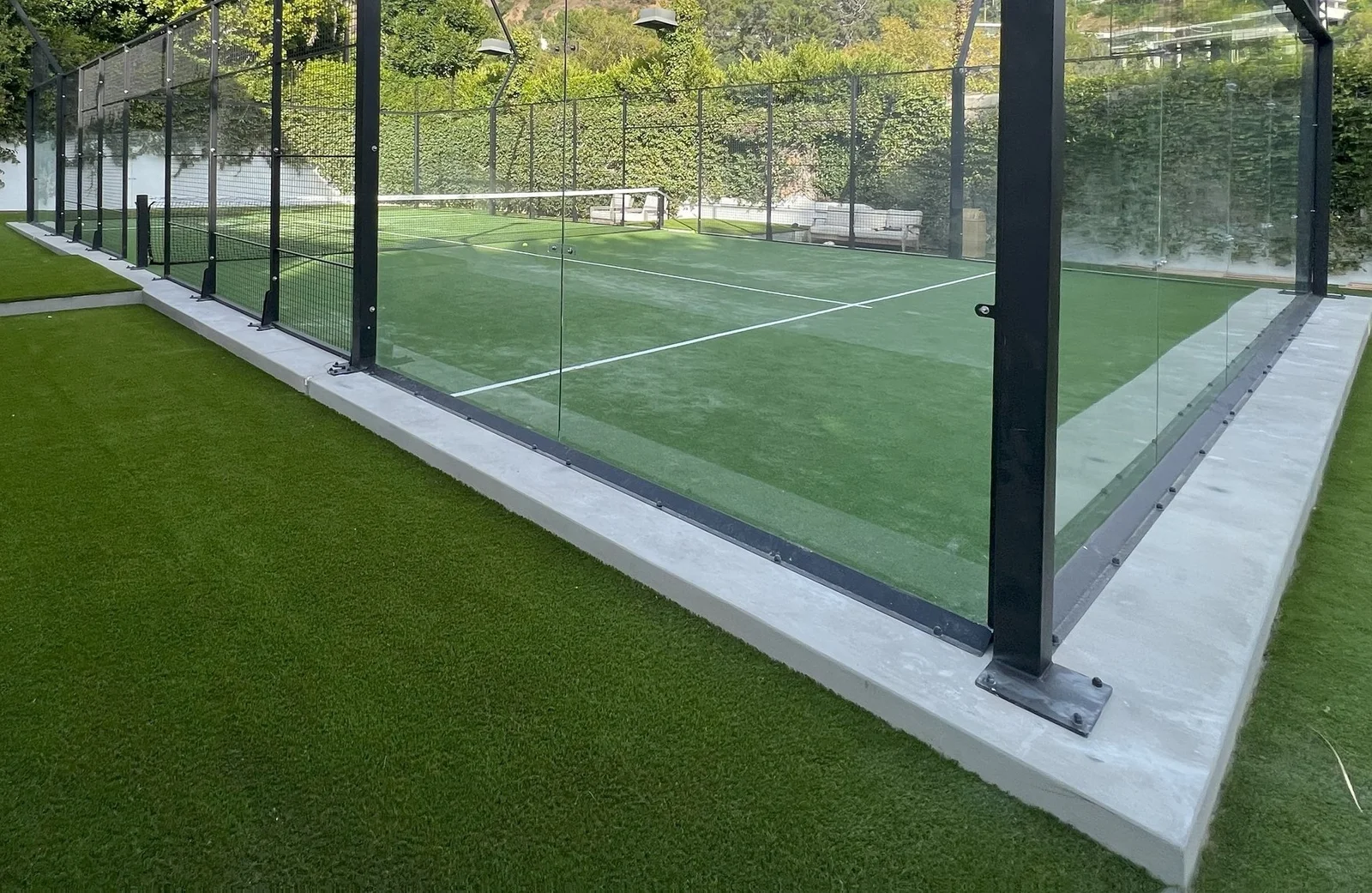 A padel court with glass walls and green artificial turf showcases expert artificial turf installation. The court is enclosed by a sleek metal and glass fence, with surrounding lush green turf. Nestled in San Tan Valley, trees and a wall complete the scenic background.