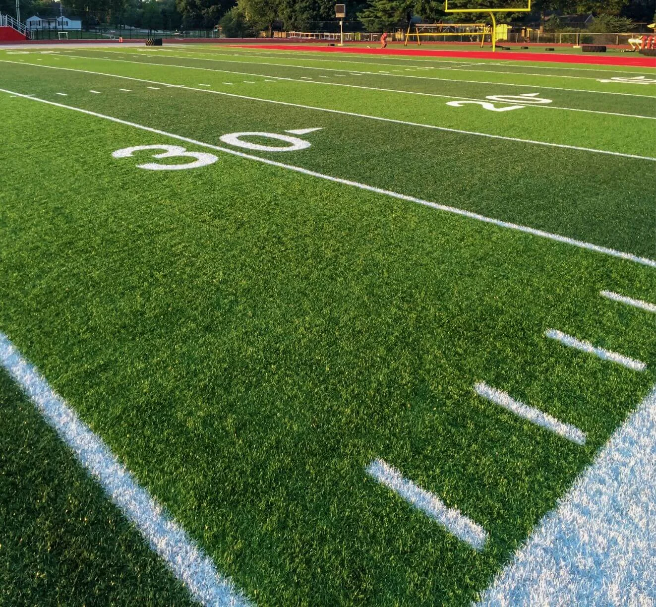 A sunlit sports field in San Tan Valley boasts vibrant green artificial turf, white yard lines, and a bright yellow goalpost in the distance. Shadows stretch across the field, hinting at late afternoon or early evening.