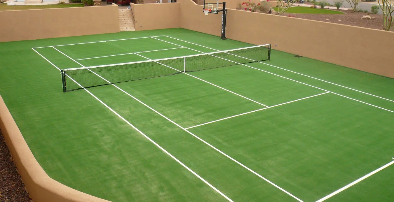 A green multi-sport court with white lines for tennis and a basketball hoop on one end showcases an impressive artificial turf installation. Surrounded by brown walls, steps lead up on the left side, with San Tan Valley's desert landscaping visible in the background.