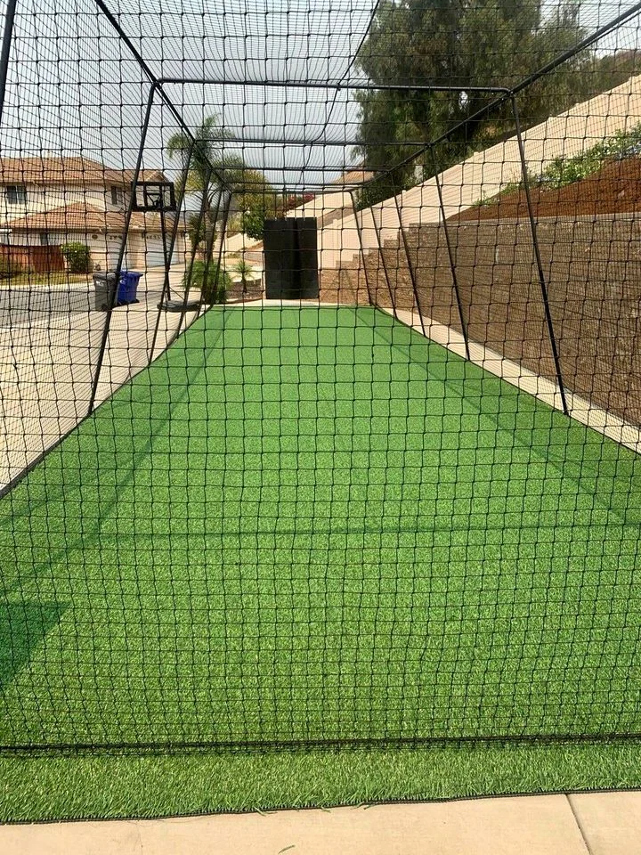 A backyard practice area in San Tan Valley featuring artificial turf installation and a tall net enclosure. The netting forms an extended rectangular cage, perfect for sports like baseball or golf. Brick walls frame the area, offering views of nearby houses.