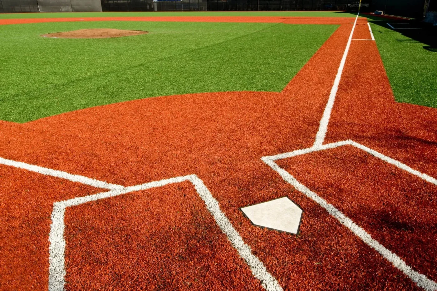 A baseball field with high-quality turf forms a geometric pattern in vibrant orange near home plate. In San Tan Valley, the pitcher's mound is visible in the distance. White lines contrast sharply against the artificial grass, marking the baselines and batter's box.