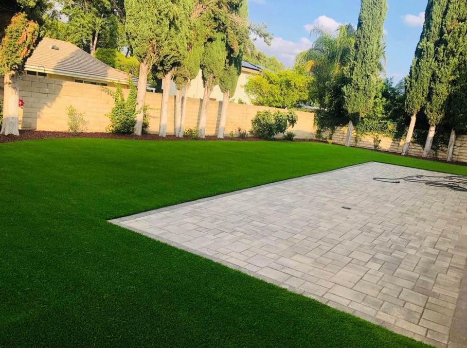 A backyard in San Tan Valley, AZ features a neatly trimmed green lawn and a paved stone patio crafted through expert paver installations. Tall trees line the stone wall fence, creating a private outdoor space. The sky is partly cloudy, enhancing the serene, well-maintained ambiance.