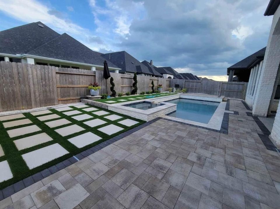 A modern outdoor space in San Tan Valley, AZ, features a swimming pool with a connected hot tub. The area is surrounded by stone tiles and a geometric grass pattern. Wooden fences and neighboring houses complete the scene under a cloudy sky, showcasing expert paver installations.