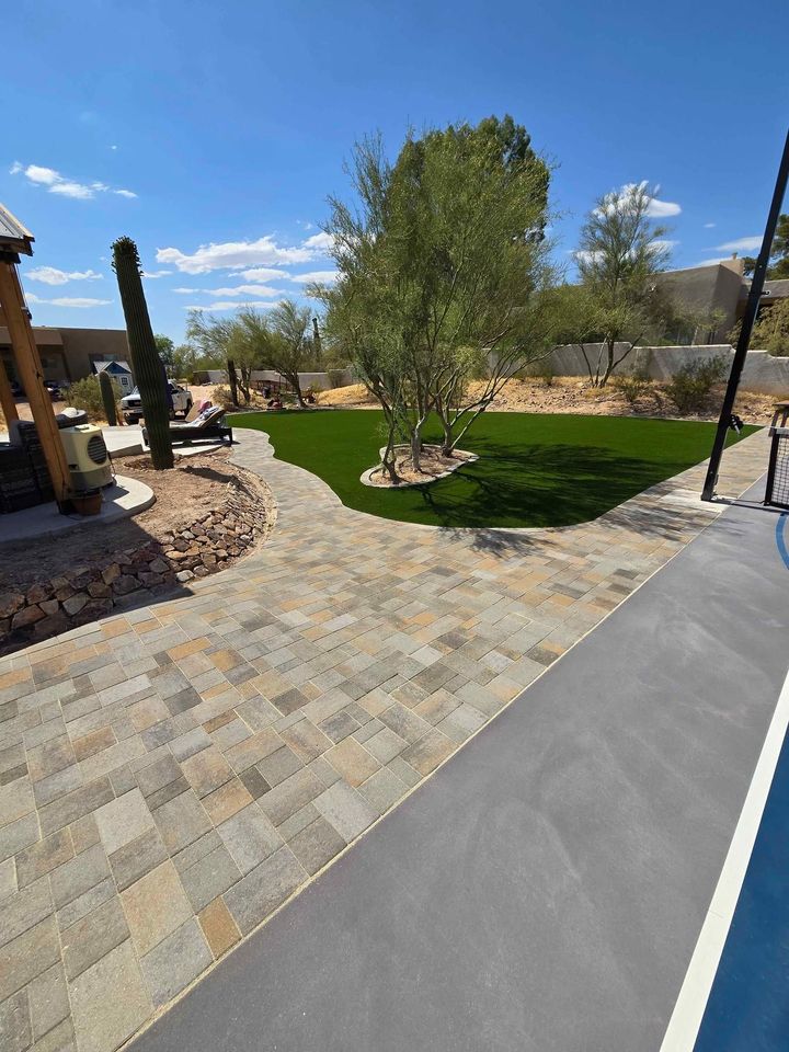 A backyard in San Tan Valley boasts a spacious stone patio, lush artificial grass lawn, and desert landscaping with cacti. The pool edge gleams on the right under a clear sky with a few clouds.