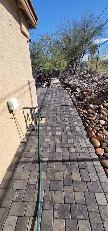 A paved walkway in this outdoor space runs alongside a building, bordered by a garden with rocks on the right. A green hose lies along the path in San Tan Valley, AZ, and trees are visible under a clear blue sky.